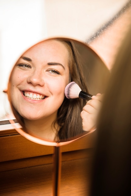 Hand mirror with reflection of happy woman applying blusher on her face