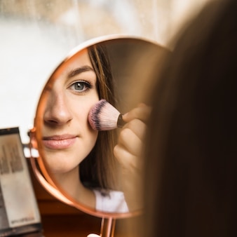 Hand mirror with reflection of woman applying blusher on her face