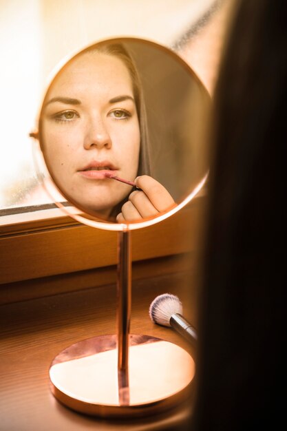 Hand mirror with reflection of woman applying lipstick on her lips