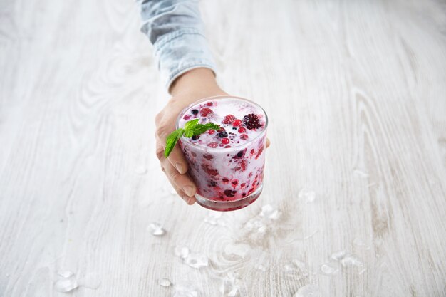 Hand offers mixed frozen berries in rox glass mixed with organic fresh milk and mint leaves on top above wooden table with melted ice cubes. Summer refreshment cocktail