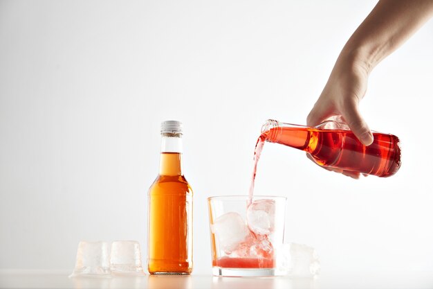 オレンジ色のアペロールが入った密封された閉じたラベルのないボトルの近くに角氷を入れたグラスにベリーサイダードリンクを手で注ぎます