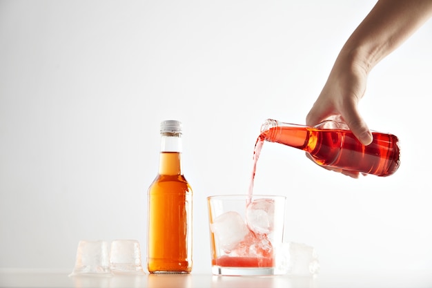 Hand pours berry cider drink into glass with ice cubes near sealed closed unlabeled bottle with orange aperol