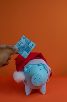 オレンジ色​の​背景​に​分離された​クリスマス​サンタクロース​の​帽子​と​貯金箱​に​本物​の​ブラジル​の​お金​を​置く​手​。​コンセプト​イメージ​。