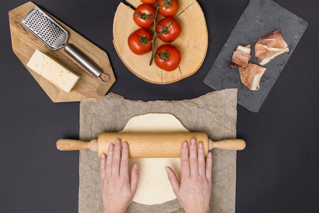 Hand rolling pizza dough on parchment paper with pizza ingredient on black background