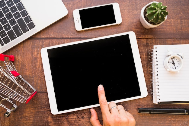 Hand on tablet near smartphone, laptop and shopping trolley