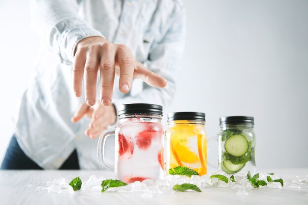 Hand takes one of rustic jars with cold fresh homemade lemonades from strawberry, orange, cucumber, ice and mint