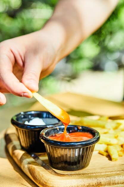 Hand taking a piece of fries with ketchup on wooden table.