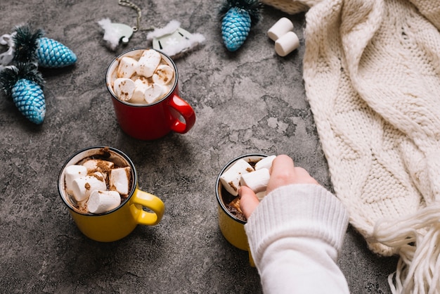 Hand with marshmallow near cup, Christmas toys and knitted scarf 