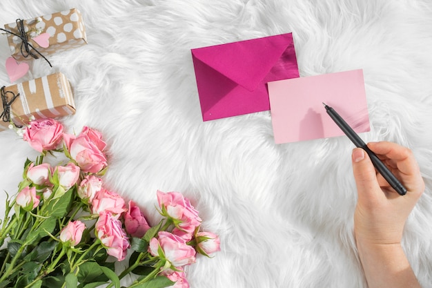 Hand with pen near paper, envelope, gifts and fresh flowers on woolen coverlet