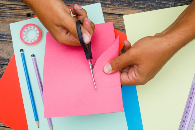 Free photo handcraft concept with tools on wooden background flat lay. man cutting heart out of paper.