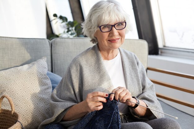 공예, 취미, 나이 및 은퇴 개념. 주름과 짧은 회색 머리를 가진 우아한 아름다운 노인 여성, 여가 시간을 즐기고, 거실에 앉아 자신을 위해 세련된 스카프 뜨개질