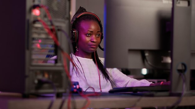 Handheld shot of african american streamer talking on headset while online playing first person shooter on gaming pc setup. Gamer girl streaming while sitting in gaming chair explaining gameplay.