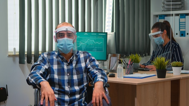 Handicapped businessman in wheelchair with mask having online conference in business office during covid-19 pandemic. Immobilized freelancer working in financial company respecting social distance.