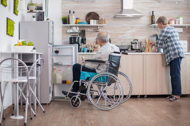 Handicapped man helping wife at the kitchen by taking eggs carton from refrigerator. Senior woman helping handicapped husband. Living with disabled person with walking disabilities