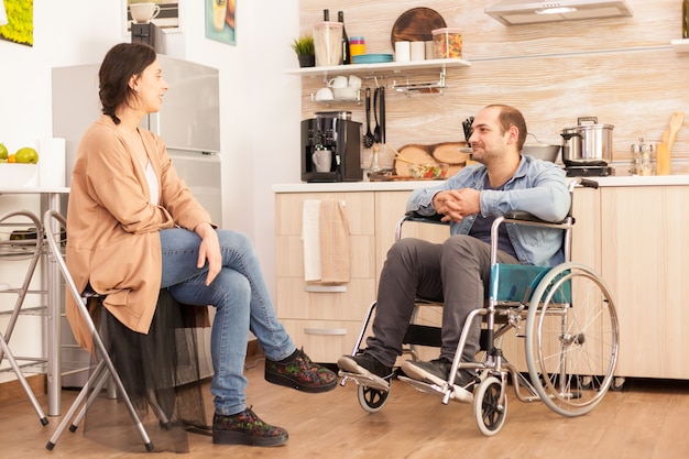 Handicapped man in wheelchair looking at caring wife in kitchen. Disabled paralyzed handicapped man with walking disability integrating after an accident.