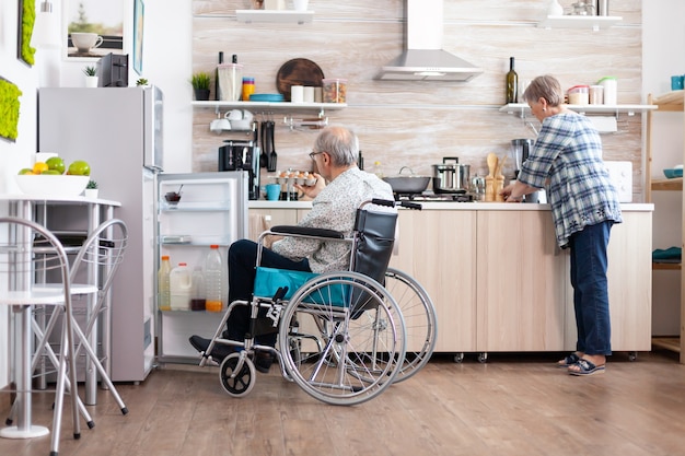 Free photo handicapped man in wheelchair opening refrigerator and helping wife preparing breakfast in kitchen. senior woman cooking for paralized husband, living with disabled man with walking disabilities