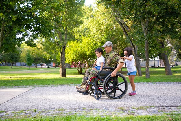Handicapped military veteran walking with two children in park. Boy sitting on dads lap, girl pushing wheelchair. Veteran of war or disability concept