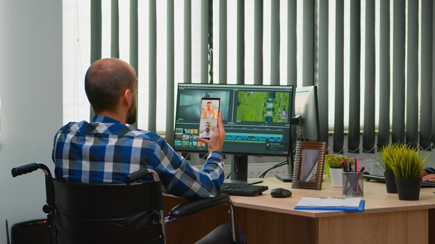 Handicapped videographer in wheelchair talking on webcam with cowerkers while editing a video project creating content in modern company office. Creator blogger working from photo studio.