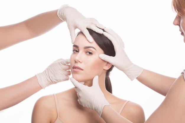 Handmade beauty Beautiful young woman keeping eyes opened while four hands in medical gloves touching her face isolated on white background