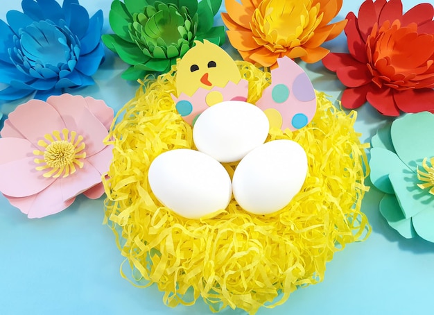 手作りの巣、さまざまな色の花、段ボールから切り取った卵の鶏。青い背景のかわいいアートの創造性。イースターでの工芸品の広告コンセプト。フラットレイ、上面図、クローズアップ、コピースペース