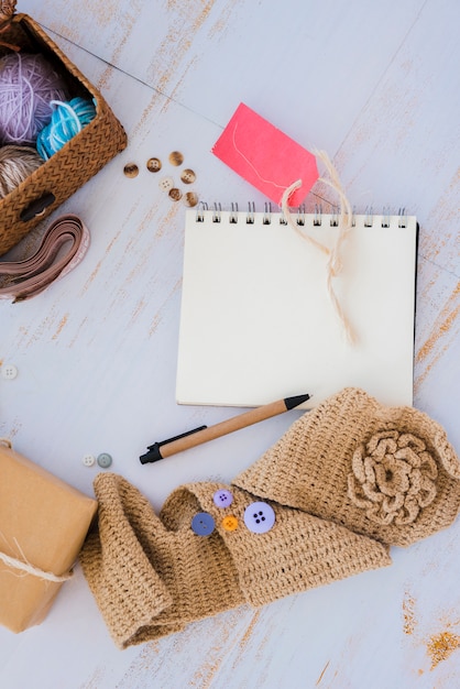 Handmade red tag; spiral notepad; pen; buttons and wool with basket on wooden desk