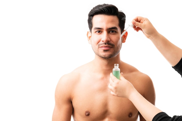 Hands of beautician applying a drop of hyaluronic acid serum on face of a young man on plain background