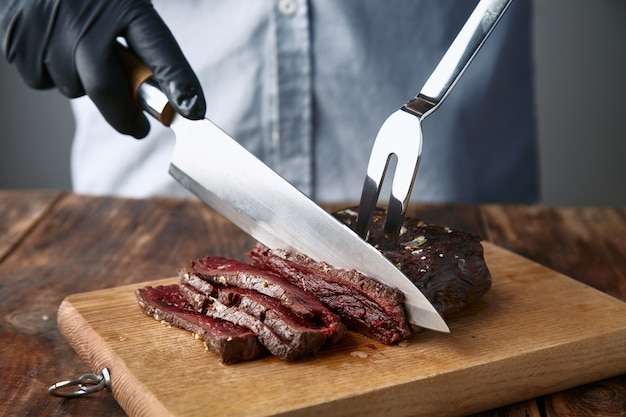 黒い手袋をはめた手は、ナイフとフォークでミディアムレアの調理済みクジラ肉ステーキをスライスします