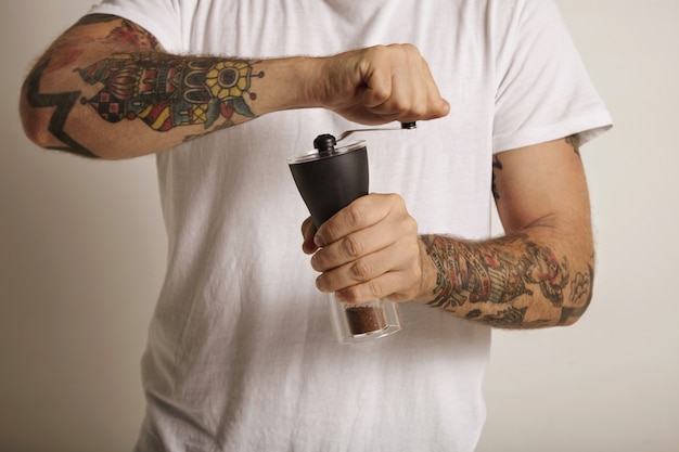 Руки и грудь молодого человека с татуировками, измельчающего кофе в ручной кофемолке