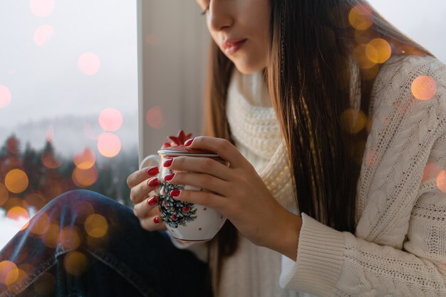 Hands close up of young attractive woman in stylish white knitted sweater sitting at home on windowsill at Christmas holding cup drinking hot tea, winter forest background view, lights bokeh