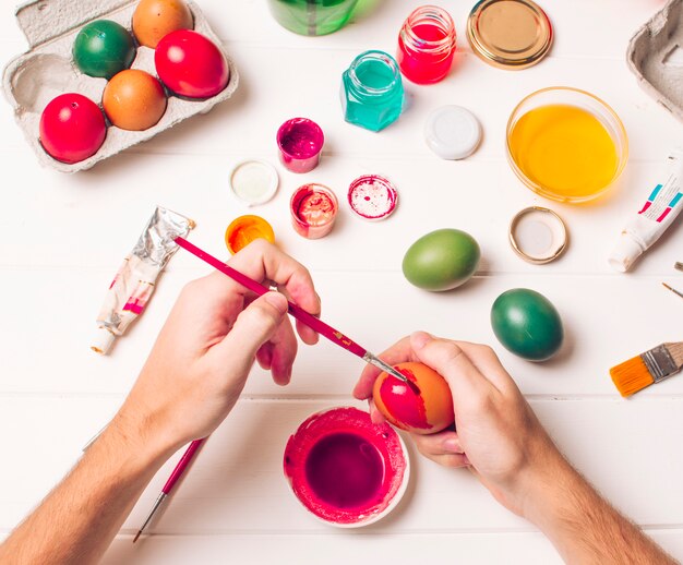 Hands coloring Easter egg near container, brushes and pink dye liquid in cans