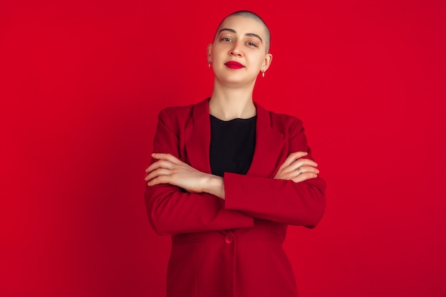 Hands crossed. Portrait of young caucasian bald woman isolated on red wall. Beautiful female model in jacket. Human emotions, facial expression, sales, ad concept. Freaky culture.