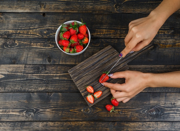 Hands cutting strawberries with knife on wooden board