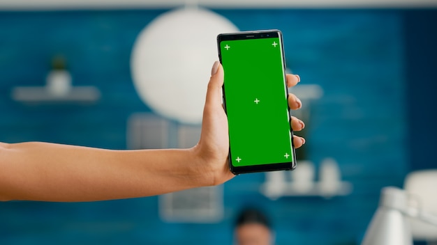 Hands of freelencer holding vertical mock up green screen chroma key smartphone. Business woman using isolated phone for browsing social networks sitting on office desk