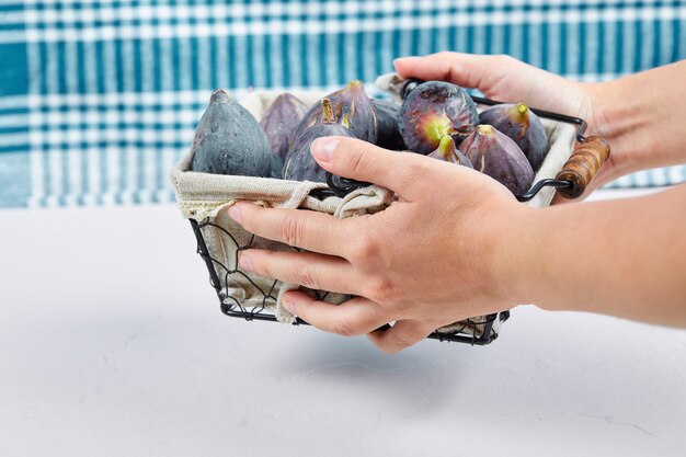 Hands holding a basket of purple figs on white with a blue tablecloth.