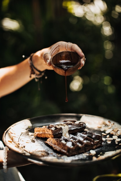 Hands holding black plate with waffles, peanuts and chocolate sauce