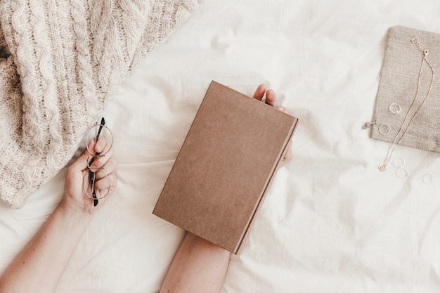 Hands holding book and eyeglasses on bed