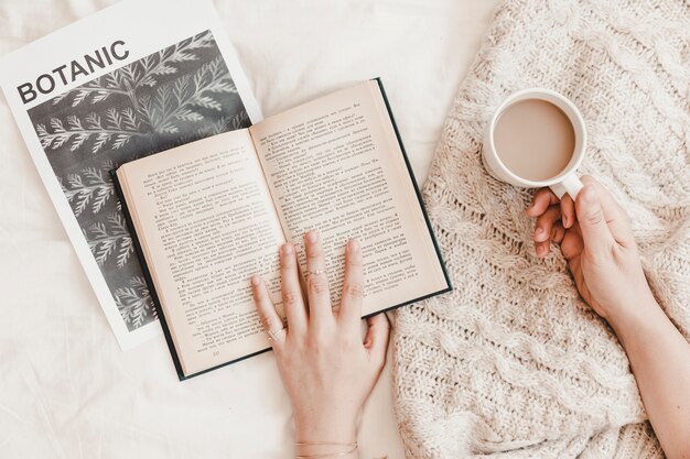 Hands holding book and hot drink lying on poster and plaid on bedsheet