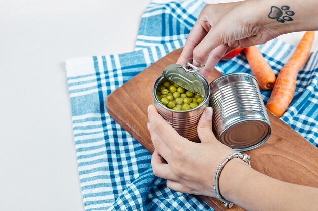 Hands holding a can of boiled green peas on a white table with vegetables and tablecloth.