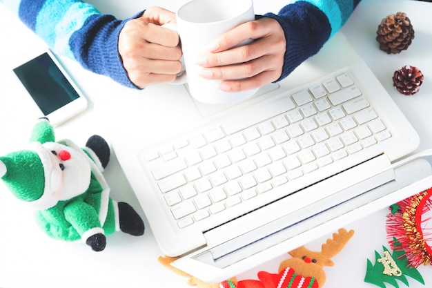 Hands holding cup of coffee and using laptop, smartphone with Christmas decoration, Shopping online