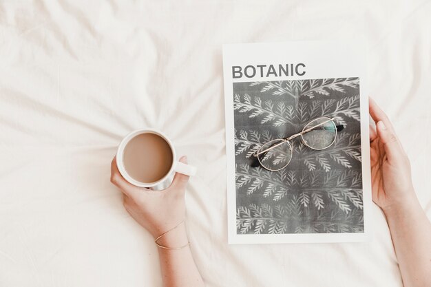 Hands holding hot drink and poster with eyeglasses on bedsheet