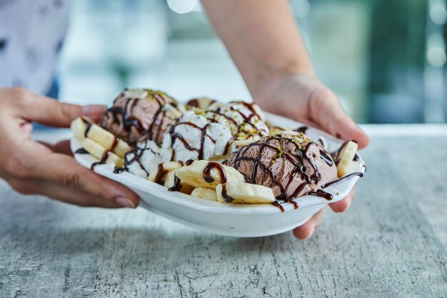 Hands holding ice-cream with bananas, chocolate, sprinkles on the white plate