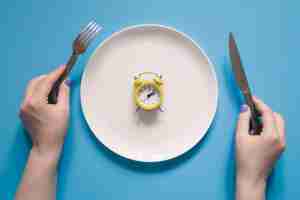 Free photo hands holding knife and fork above alarm clock on a plate on blue background