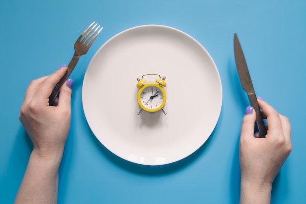파란색 배경에 있는 접시에 알람 시계 위에 나이프와 포크를 들고 있는 손