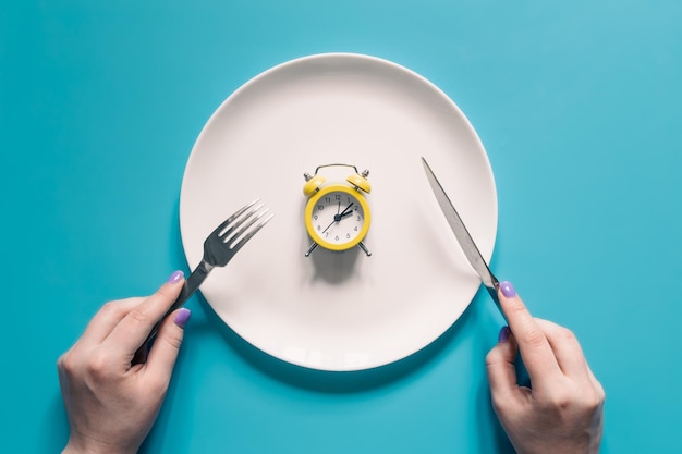 Free photo hands holding knife and fork above alarm clock on a plate on blue background