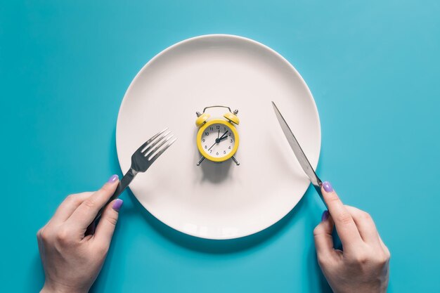 Hands holding knife and fork above alarm clock on a plate on blue background