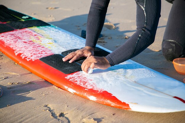 Руки мужчины-серфера в гидрокостюме, смазывая доску для серфинга на песке на берегу океана