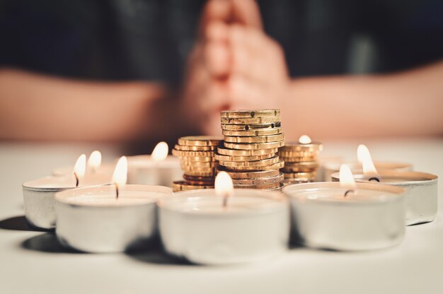 Hands of a man praying with a circle of burning candles with a stack of coins inside