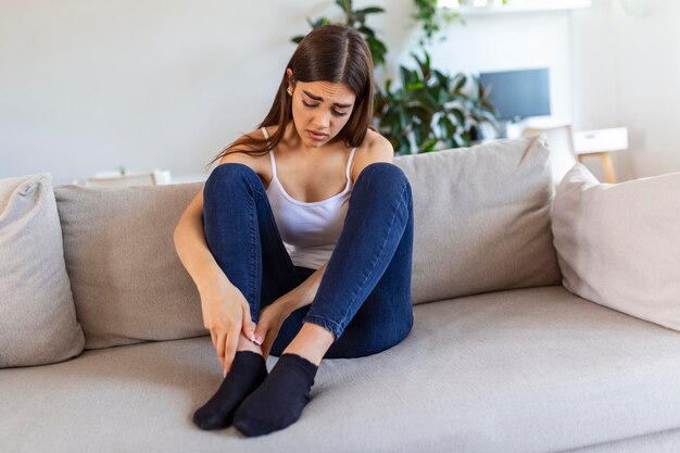 Hands massaging swollen foot while sitting on sofa during the day at home Photo of Young Caucasian woman suffering from pain in leg Woman massaging her legs after all day at work in office