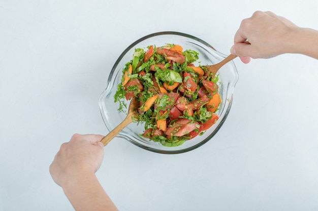 Руки, смешивающие вкусный овощной салат на стеклянной тарелке.