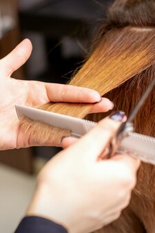 Руки парикмахера держат прядь волос между пальцами, делая стрижку длинных волос молодой женщины расческой и ножницами в парикмахерской крупным планом